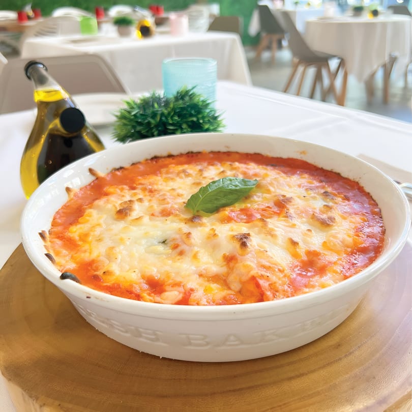 Baked pasta with melted cheese and tomato sauce, garnished with basil, on a wooden board.