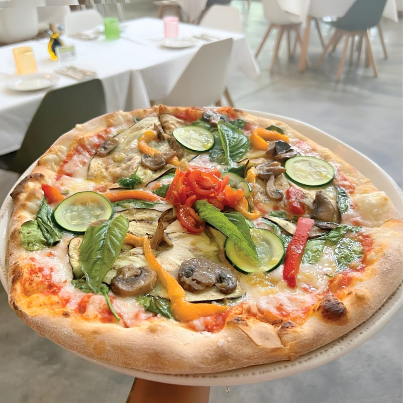 A vibrant vegetarian pizza with zucchini, mushrooms, bell peppers, and basil, held in a restaurant.